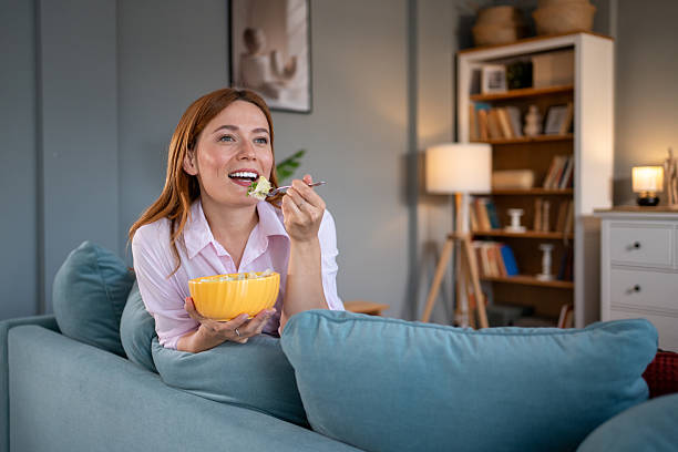 istock Smiling woman eating salad while watching tv on sofa at home 2217021799