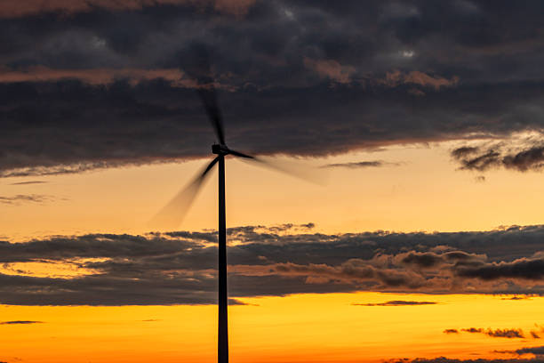 A towering wind turbine stands silhouetted against a golden-orange sunset, framed by dramatic clouds in the sky. The scene evokes sustainable energy and natural beauty in the evening light.