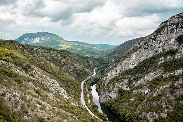 Picturesque landscape featuring a river winding through a lush green valley surrounded by majestic mountains and a scenic roadway under a partly cloudy sky.