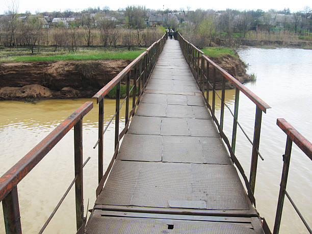 Rustic metal bridge spans a muddy river in a rural landscape. Represents connection, transition, journey. Suitable for themes of travel, resilience, or overcoming obstacles.