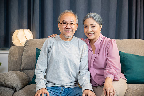 istock Happy Asian senior couple greeting someone while sitting on a comfortable couch, old man and woman sit close together on a sofa 2217073897