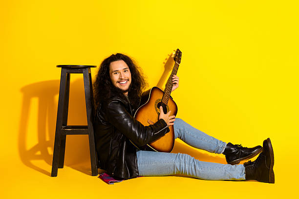 istock Young man in a black leather jacket playing guitar on a vibrant yellow background, exuding creativity and musical energy 2217083050