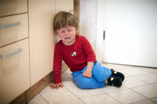 A young child in a red shirt and blue pants sits on the kitchen floor next to the cabinets, visibly upset, with tears in their eyes, conveying a strong emotional response.