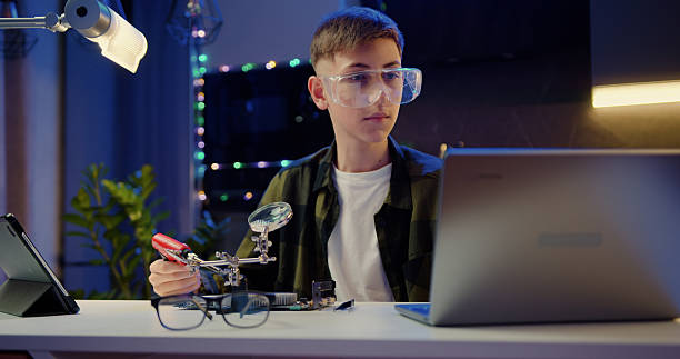 istock Smart teenage schoolboy in protective glasses soldering panel using soldering iron during revision training video on laptop in the evening at home 2217096152
