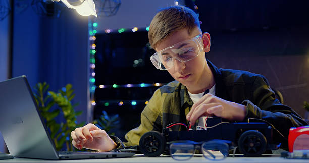 istock Smart teenage schoolboy using laptop and carefully disassembling his remote controlled toy car homework project at home. People with futeristic technology and education concept. 2217098347