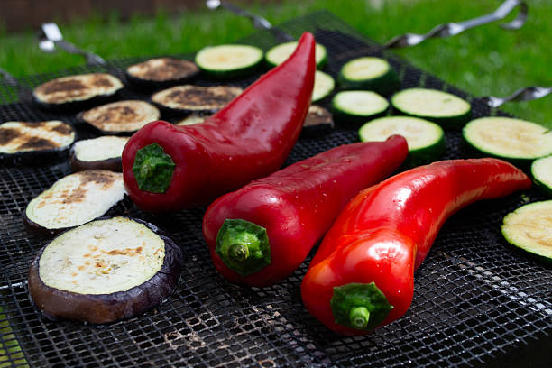 Close-up of vibrant red peppers, eggplant, and zucchini slices being grilled on a barbecue outdoors.
