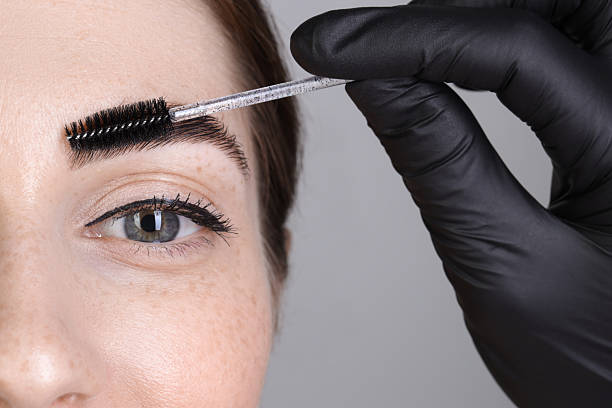 istock Brow lamination. Cosmetologist combing woman's eyebrows with brush against grey background, closeup 2217118364