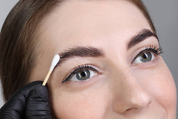 istock Brow lamination. Cosmetologist applying cream onto woman's eyebrows against grey background, closeup 2217118481