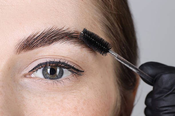 istock Brow lamination. Cosmetologist combing woman's eyebrows with brush against grey background, closeup 2217118508