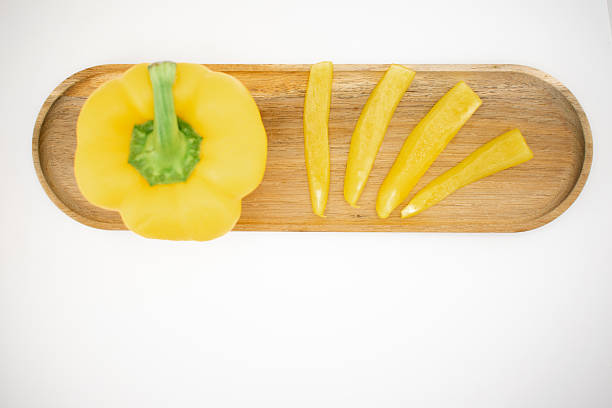 Yellow bell pepper (Capsicum annuum) sliced and arranged on a wooden surface, simulating a sun with radiating rays. The pepper's vibrant yellow hue contrasts with the natural wood and the white background. The arrangement creatively highlights the pepper's smooth, glossy texture, showcasing its freshness and potential as a culinary ingredient.