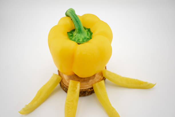 Yellow bell pepper (Capsicum annuum) sliced and arranged on a wooden surface, simulating a sun with radiating rays. The pepper's vibrant yellow hue contrasts with the natural wood and the white background. The arrangement creatively highlights the pepper's smooth, glossy texture, showcasing its freshness and potential as a culinary ingredient.