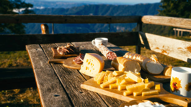 A Rustic Wooden Table is Set with a Spread of Sliced Cheese,Salami,and Fresh Bread During an Outdoor Picnic in the Mountains. The Scene is Bathed in Warm Sunlight with a Scenic View of Distant Hills and Valleys in the Background. The Arrangement Suggests a Casual Alpine Meal Shared in Nature,Evoking Themes of Travel,Food Culture,and Traditional Cuisine