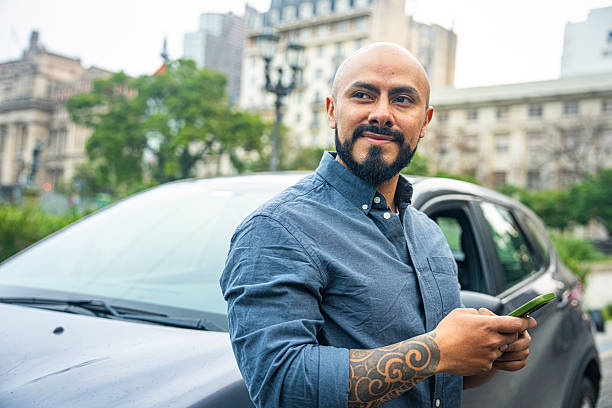 Mid-shot side view of relaxed Latin American man leaning on car and checking messages on mobile phone while waiting for friend.