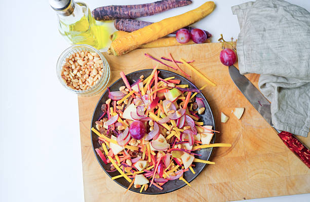 Directly above view of healthy and delicious salad with yellow and purple carrots, red grapes, apple, decorated with pine nuts and raisins on steel gray plate on  wooden cutting board, ready to be served. Photo taken with Nikon Z7ii and Nikon Z 35mm f/1.8 S lens