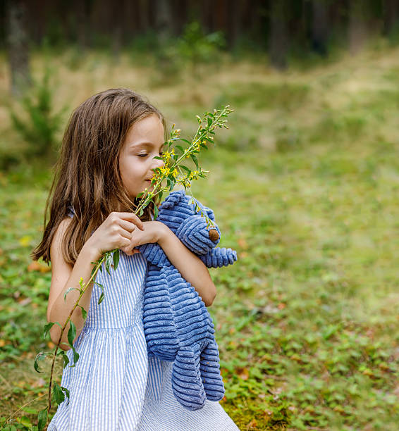 Little girl in a striped dress standing in a peaceful forest, gently holding a blue stuffed bear while smelling a delicate flower, evoking innocence and wonder