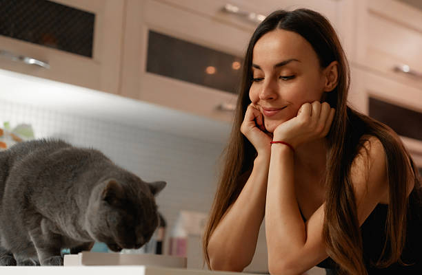 A woman smiles gently while watching her gray cat, who is sniffing something on a kitchen counter. The cozy kitchen background adds warmth to the scene, capturing a moment of connection.