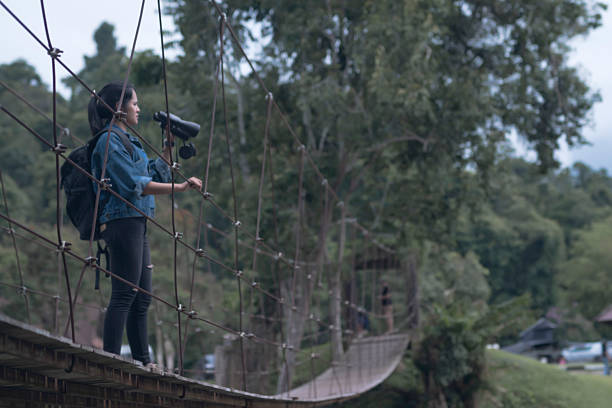 woman standing   using binoculars for birdwatching. She wears a camouflage shirt layered with a denim jacket and appears focused on observing birds in the distance.