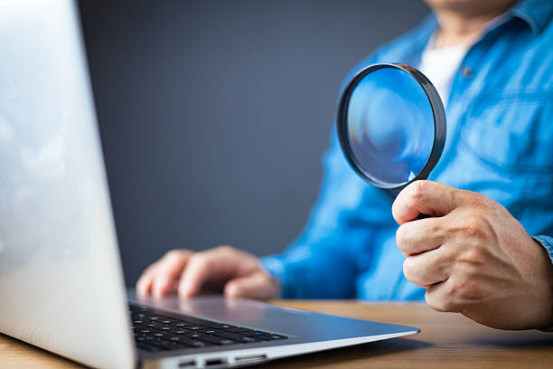 istock A man in a blue shirt holds a magnifying glass while using a laptop, symbolizing online research, investigation, cybersecurity, data analysis, or scrutiny in digital environments on a wooden desk. 2217138658