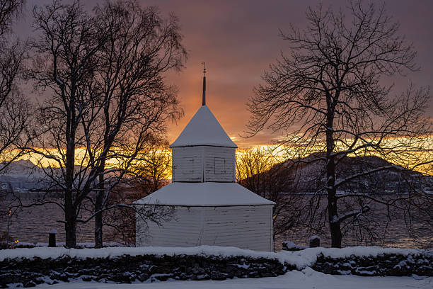 White wooden chapel, surrounded by cemetery and stone fence in the arctic autumn with snow. Norwegian sea and mountains in the background. Colorful sky during sunrise, Harstad, Norway.