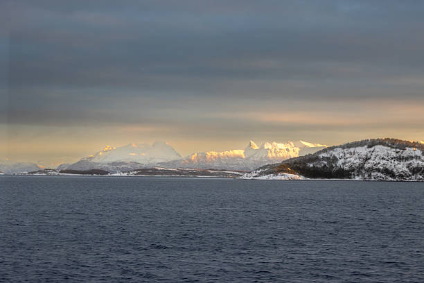 Calm water of Norwegian sea. Mountains covered by snow, bright yellow color in the arctic sunrise. Colorful sky. Harstad, Norway.
