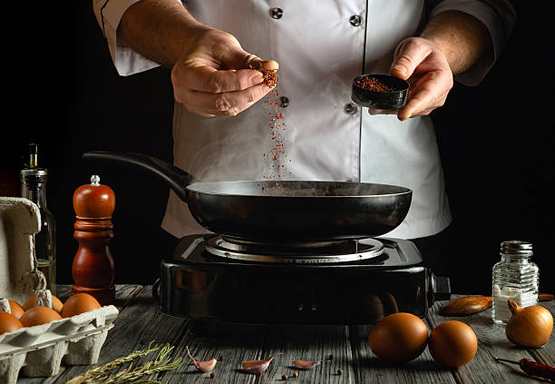 istock A skilled chef is sprinkling spices into a frying pan on a stove, surrounded by fresh eggs, seasoning jars, and kitchen tools, focused on creating a delicious meal 2217149040