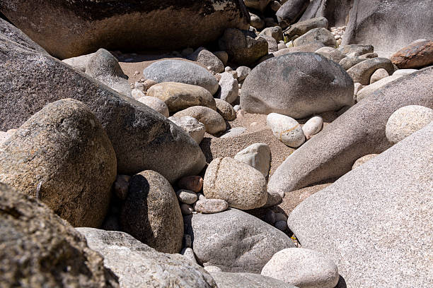 High-angle, close-up shot of a collection of various-sized rounded rocks and stones.  The rocks exhibit a range of grey and light grey hues, with variations in texture, showing both smooth and rough surfaces.  The image displays strong contrasts between light and shadow created by natural sunlight illuminating the uneven rock surfaces. The overall composition focuses on the textural details and natural color palette of the stones.  Depth of field is shallow, keeping the focus primarily on the rocks in the foreground.