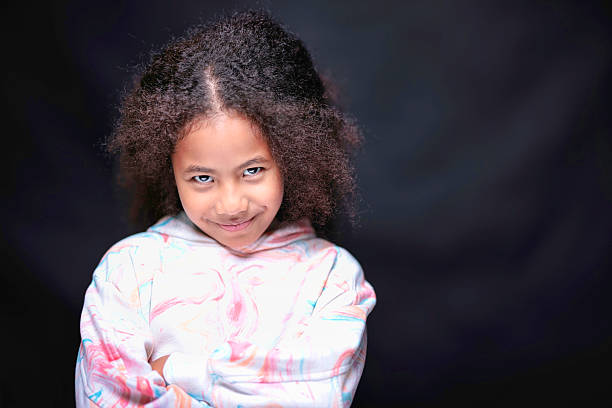 Cute little girl with blonde hair and blue eyes, dressed with a blue navy dress, holds her hands together, smiling she's looking at the camera, the background is blacked, two speedlights were used for the shot.