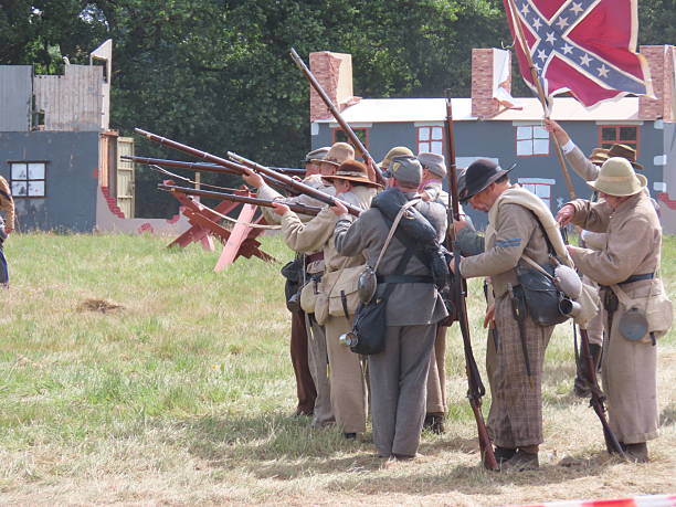 A collection of medieval swords displayed on a wooden rack at an outdoor historical reenactment event. The swords feature various hilt designs, including a leather-wrapped grip and a crossguard, reflecting traditional craftsmanship. The background includes a medieval-style tent and wooden furniture.