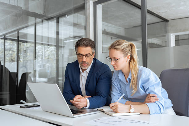 istock Two partners, team of professional business people working, discussing, writing notes at desk. Mature focused senior mentor leader man showing young woman manager project on laptop in modern office 2217161478