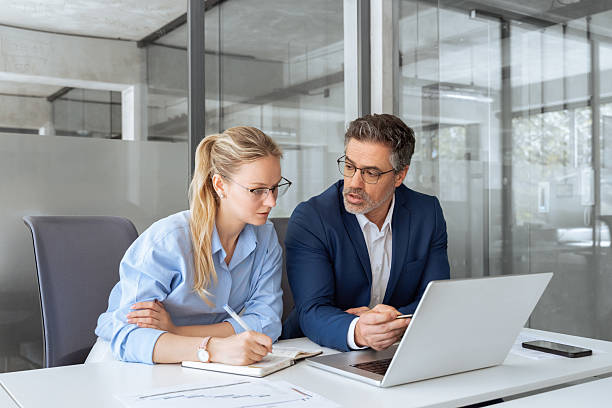 istock Two partners, team of professional business people working, studying, writing notes sit at desk. Mature focused senior mentor leader man showing young woman manager project on laptop in modern office 2217161479