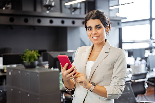istock Young Latina professional woman in blazer using smartphone mobile at office, looking at camera. Portrait of smiling businesswoman in modern workspace, holding cellphone for work chat or business call 2217163644