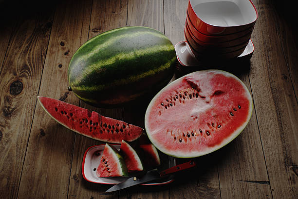 Digitally rendered rustic wooden table adorned with a whole watermelon and neatly sliced pieces. A sharp kitchen knife is accompanied by red and white bowls, enhancing the presentation. The image captures the refreshing essence of watermelon, perfect for summer moments or culinary themes.