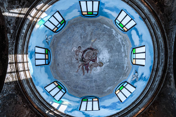 Looking up into the decaying dome of an abandoned church, a faded fresco of angels amidst a painted sky is illuminated by light streaming through weathered windows