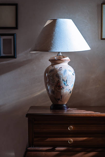 An elegant still life of a vintage, oriental-style ceramic table lamp decorated with butterflies and flowers. Resting on a classic wooden console table, the lamp is artfully lit, with dramatic light and shadows playing on the wall behind it, creating a warm, timeless, and sophisticated atmosphere