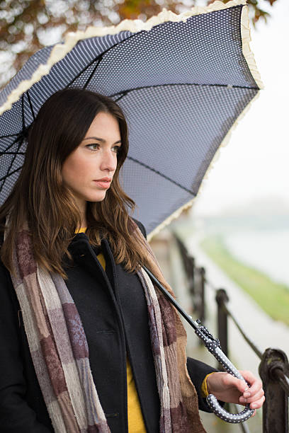 Woman in a striped T-shirt under a black umbrella standing in the rain in a green city courtyard. Dark-toned moody image