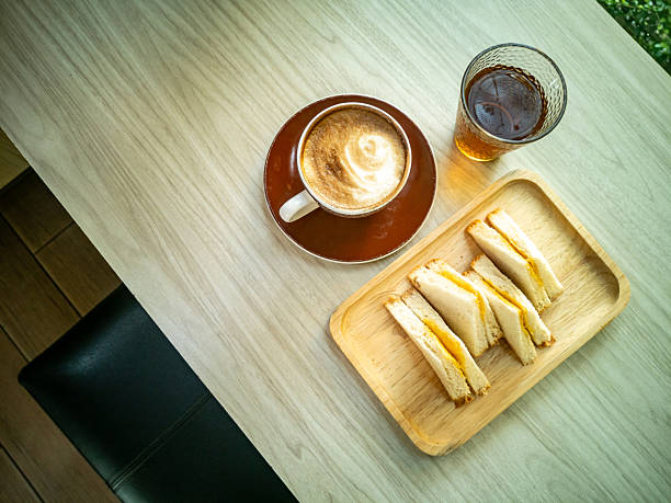 A cozy breakfast scene featuring a cup of coffee with latte art, a glass of cold beverage, and a plate of fresh sandwiches on a wooden tray.