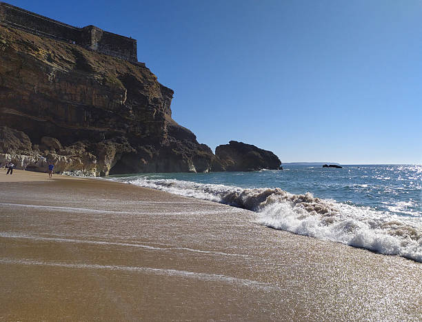 istock Scenic beach view featuring golden sand, rolling waves, and dramatic cliffs topped with historic buildings under a clear blue sky. Ideal for travel, vacation, or coastal themes. 2217224550