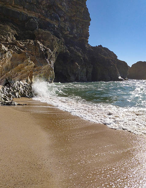 Scenic ocean shore featuring a rocky cliff face, sandy beach, and breaking wave. Evokes tranquility, travel, and the power of nature. Ideal for vacation or coastal themed projects.