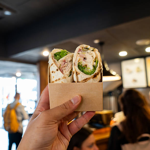 istock A Hand Holding a Takeaway Chicken Caesar Salad Wrap in Front of a Bustling Cafe Counter Showcases Fresh Grab and Go Dining Inside a Trendy London Eatery. 2217228805
