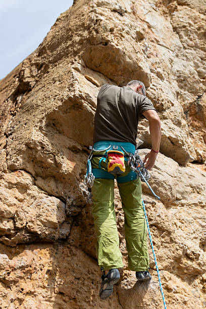 A man is climbing a rock wall with a blue and green backpack. He is wearing a brown shirt and green pants