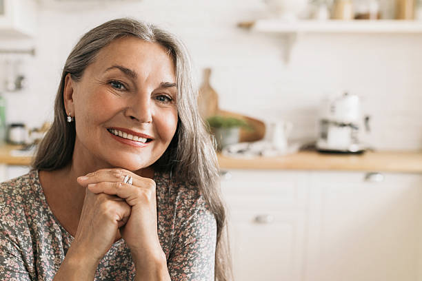 istock Smiling elderly female sitting at kitchen table. Cheerful middle-aged woman looking at camera with happiness and calmness. Aging and mental balance concept 2217243890