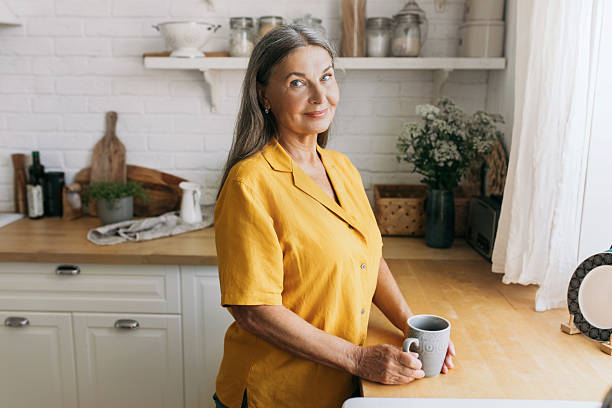 istock Charming senior lady enjoying view from window. Cozy kitchen interior with fresh flower. Wake up in countryhouse, weekends away from city, slow morning routine, coffee time concept 2217243899