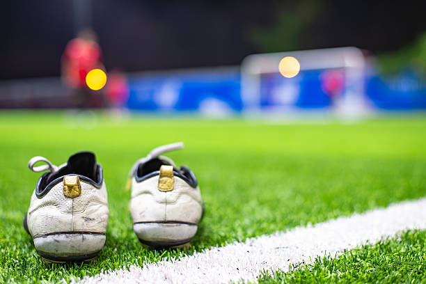 Worn soccer cleats rest on artificial turf as blurred players warm up near the goalpost during a nighttime practice session.