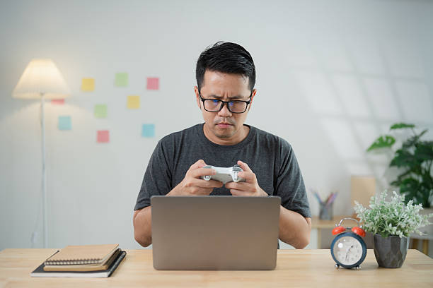 istock Young man with glasses focused on gaming controller while sitting at desk with laptop in modern workspace with plants and soft lighting 2217251471