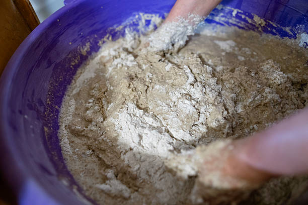 istock Hands kneading dough in a large plastic container during the process of preparing homemade bread. The dough is sticky and textured, showing active mixing. 2217268499