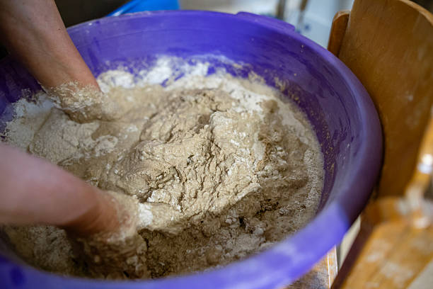 istock Hands kneading dough in a large plastic container during the process of preparing homemade bread. The dough is sticky and textured, showing active mixing. 2217268627