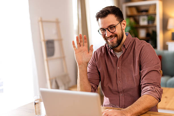 istock Friendly man greeting someone on a video call while working from home. He sits in a stylish kitchen with a laptop, orange juice, and warm lighting, creating a welcoming and professional setting. 2217281124