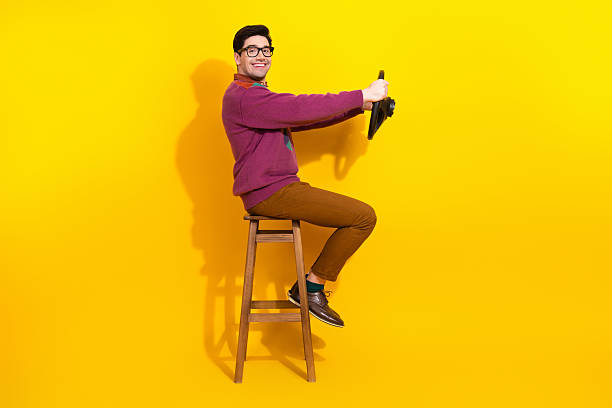istock Young man with glasses performing a humorous driving gesture on a stool against a vibrant yellow background 2217282447
