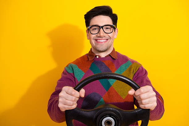 A cheerful young man holding a steering wheel with both hands, smiling enthusiastically against a bright yellow background, embodying positivity and retro charm.