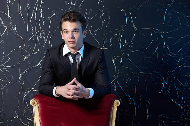 Attractive young man dressed in business style stands behind a red chair, portrait on a black textured background, studio shooting